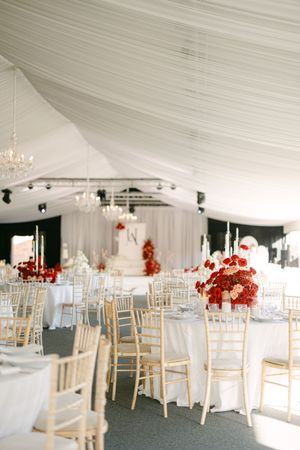 Elegant wedding reception tent with white draping, crystal chandeliers, gold chairs and red floral centerpieces.