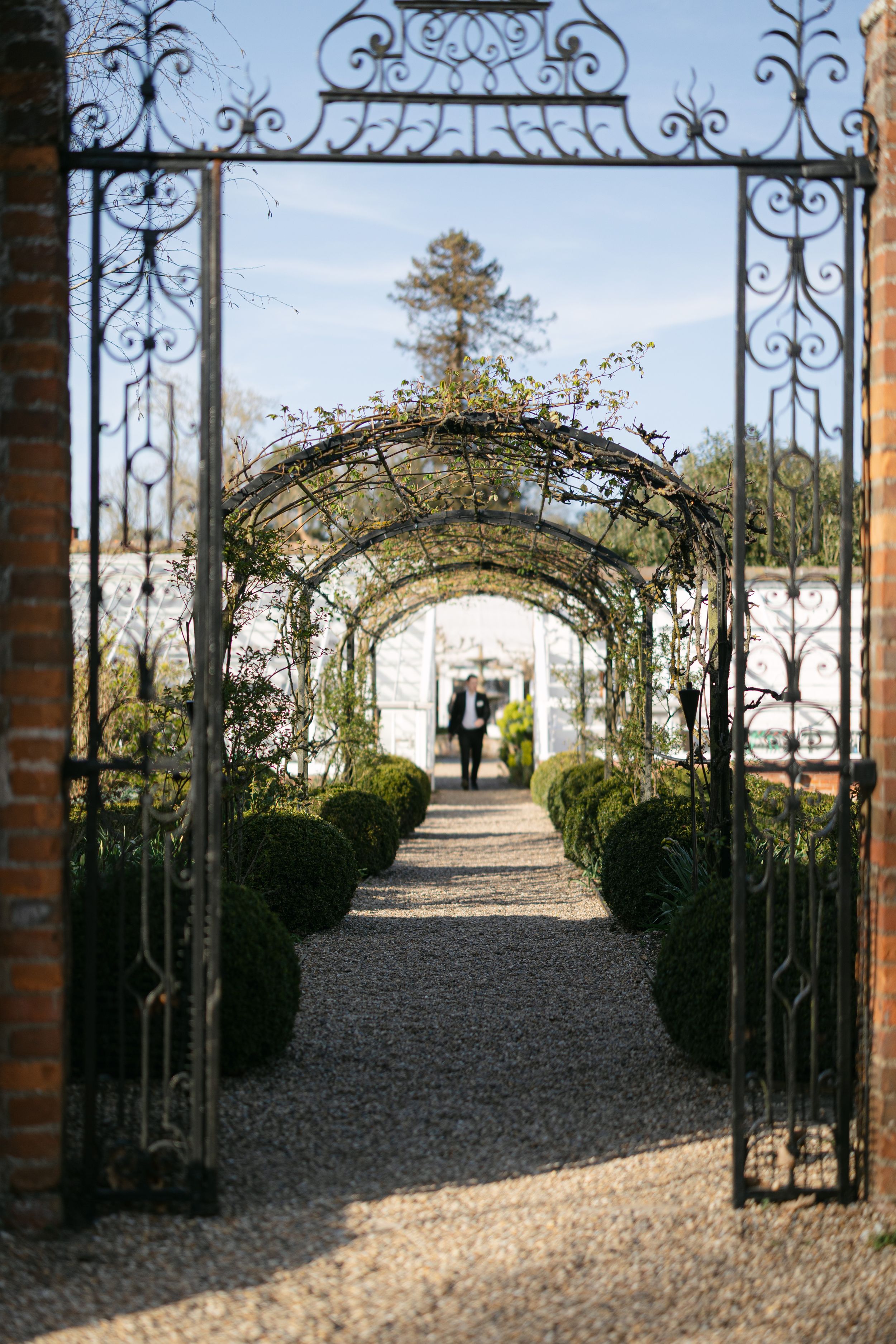 Ornate iron gates open to a garden path lined with manicured hedges leading to a distant estate.
