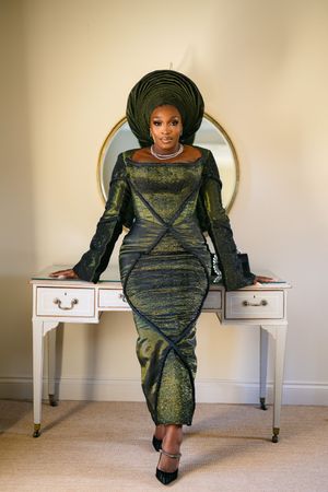 Woman in traditional African attire with gele headwrap poses elegantly on white vanity desk wearing metallic green dress.