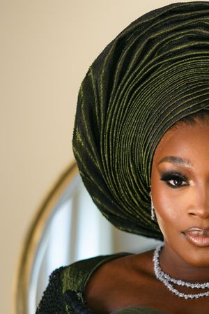 A close-up portrait featuring an elegant green gele headwrap and sparkling diamond necklace against a light background.