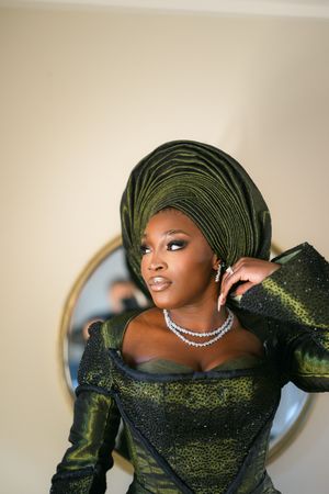 Elegant woman in emerald green traditional gele headwrap and formal dress with diamond necklace against neutral background.