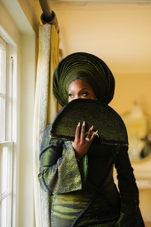Person in emerald green traditional Nigerian attire with gele headwrap poses elegantly by window in natural light.