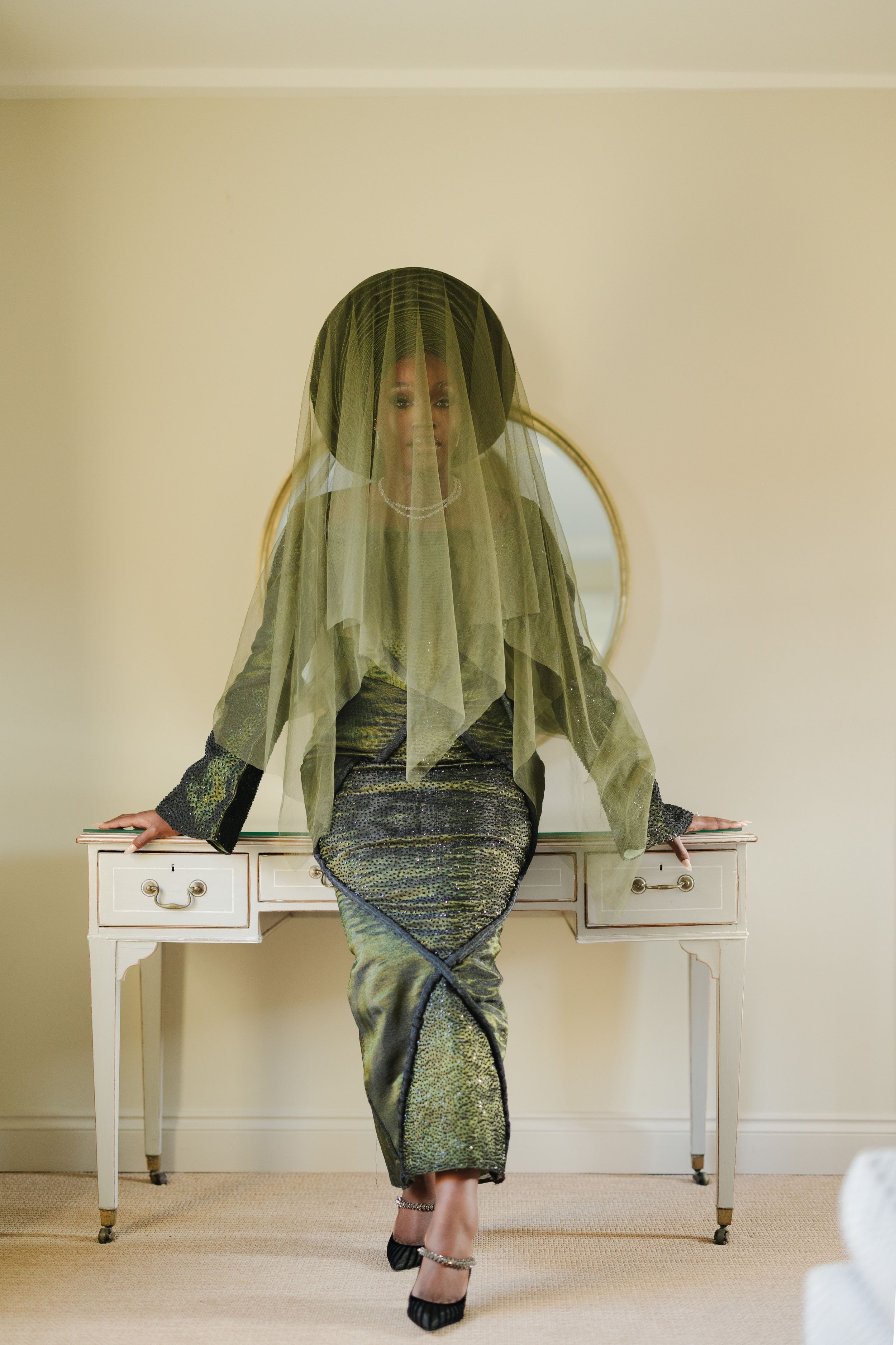 Figure in sage green veil sits atop white vintage desk wearing metallic dress and black heels.