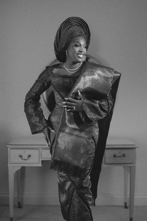 A black and white photo of someone wearing an elegant African-style dress and headwrap posing next to a desk.