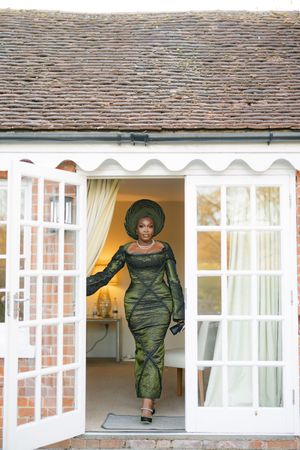 Traditional African formal dress in dark green shown stepping through French doors of a countryside home.