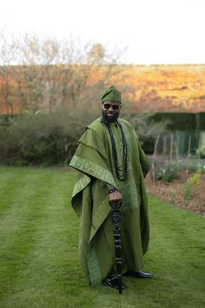 Figure in an olive green cape and sunglasses poses outdoors on grass during golden hour.