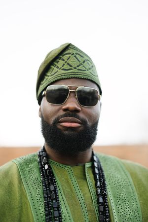 Portrait of person wearing green traditional African attire with sunglasses and beaded necklaces against white background.
