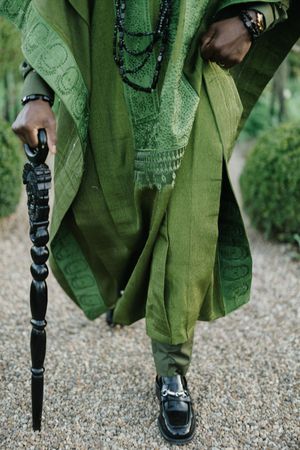 Detail shot of ornate green agbada robe and decorative walking stick against gravel path.