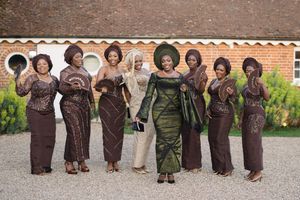 Wedding party wearing coordinated brown and olive green traditional formal attire standing in front of brick wall.