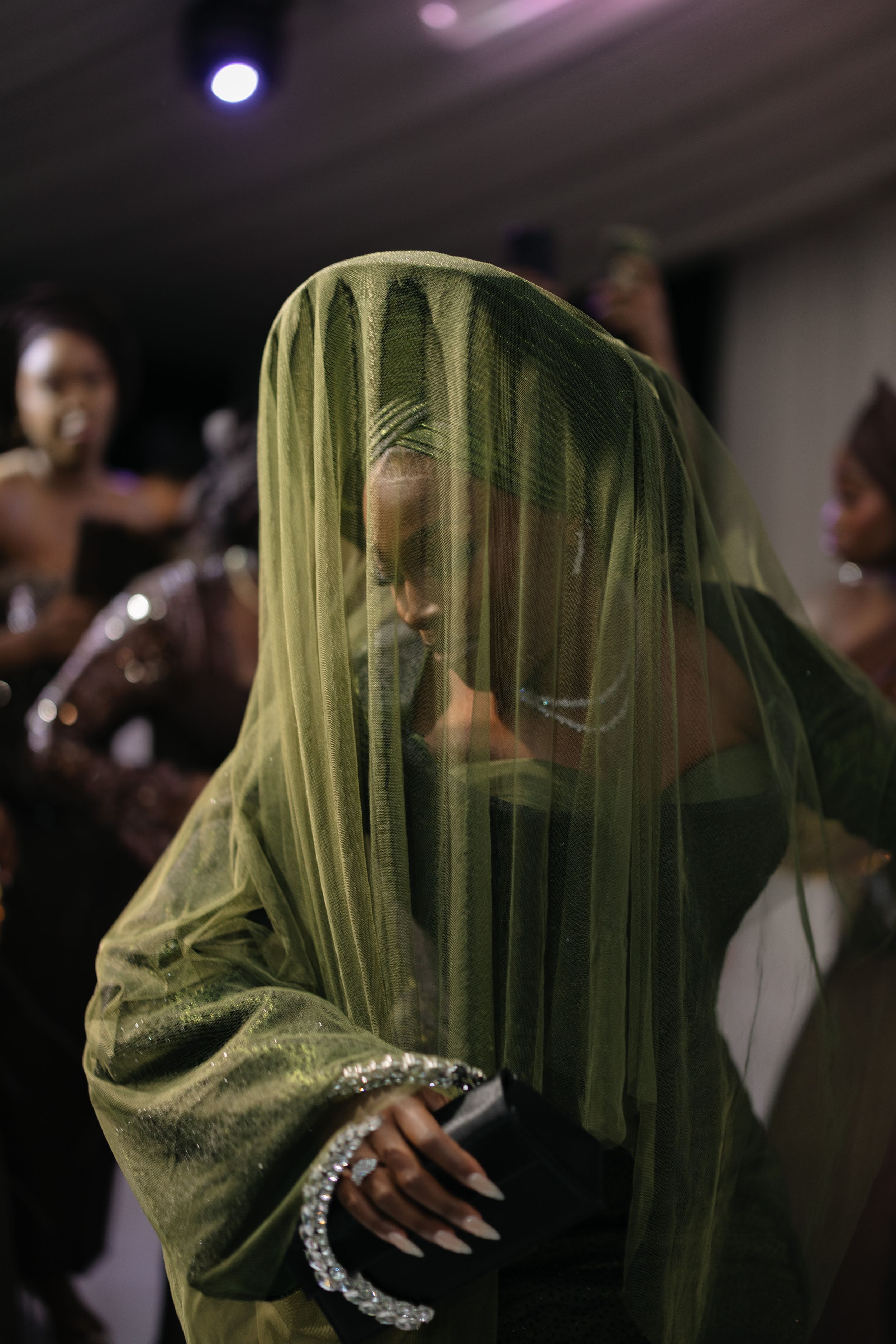 Figure on stage draped in translucent lime green fabric with silver bracelet performs under dramatic lighting.
