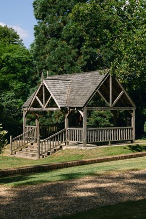 Wooden gazebo with peaked roof and railings in a peaceful garden setting.