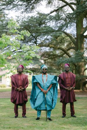 Three men in traditional African attire, with center figure in turquoise and others in burgundy, standing outdoors.