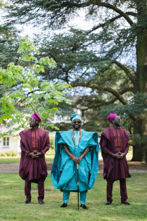 Three figures pose in traditional West African attire with burgundy and turquoise agbada robes in an outdoor garden setting.
