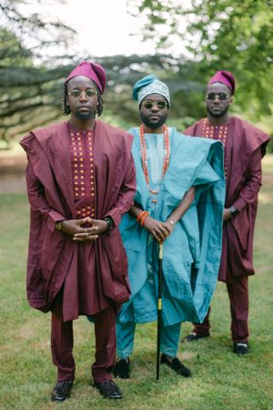 Three people in traditional African dress stand together wearing burgundy and turquoise agbada with beaded details.