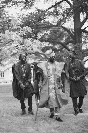 Black and white photo shows three people in traditional African formal wear walking outdoors.