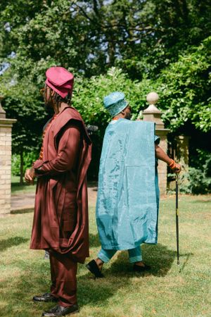 Two figures in traditional African attire, one in burgundy and one in turquoise, walking through a garden setting.