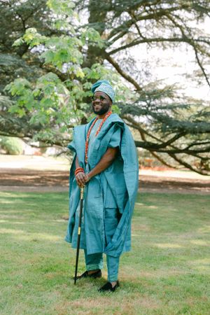 Person wearing a flowing turquoise traditional African agbada and hat standing with a walking stick in a garden.