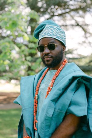 Traditional African fashion featuring turquoise agbada, patterned cap, coral beads and sunglasses.