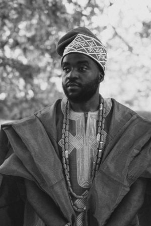 Portrait in traditional African attire featuring geometric patterned cap and embroidered robe in monochrome.