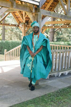 Figure in turquoise traditional African agbada seated under wooden pavilion structure.
