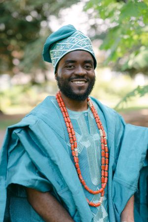 Person wearing vibrant turquoise traditional African attire with orange beaded necklaces in an outdoor setting.