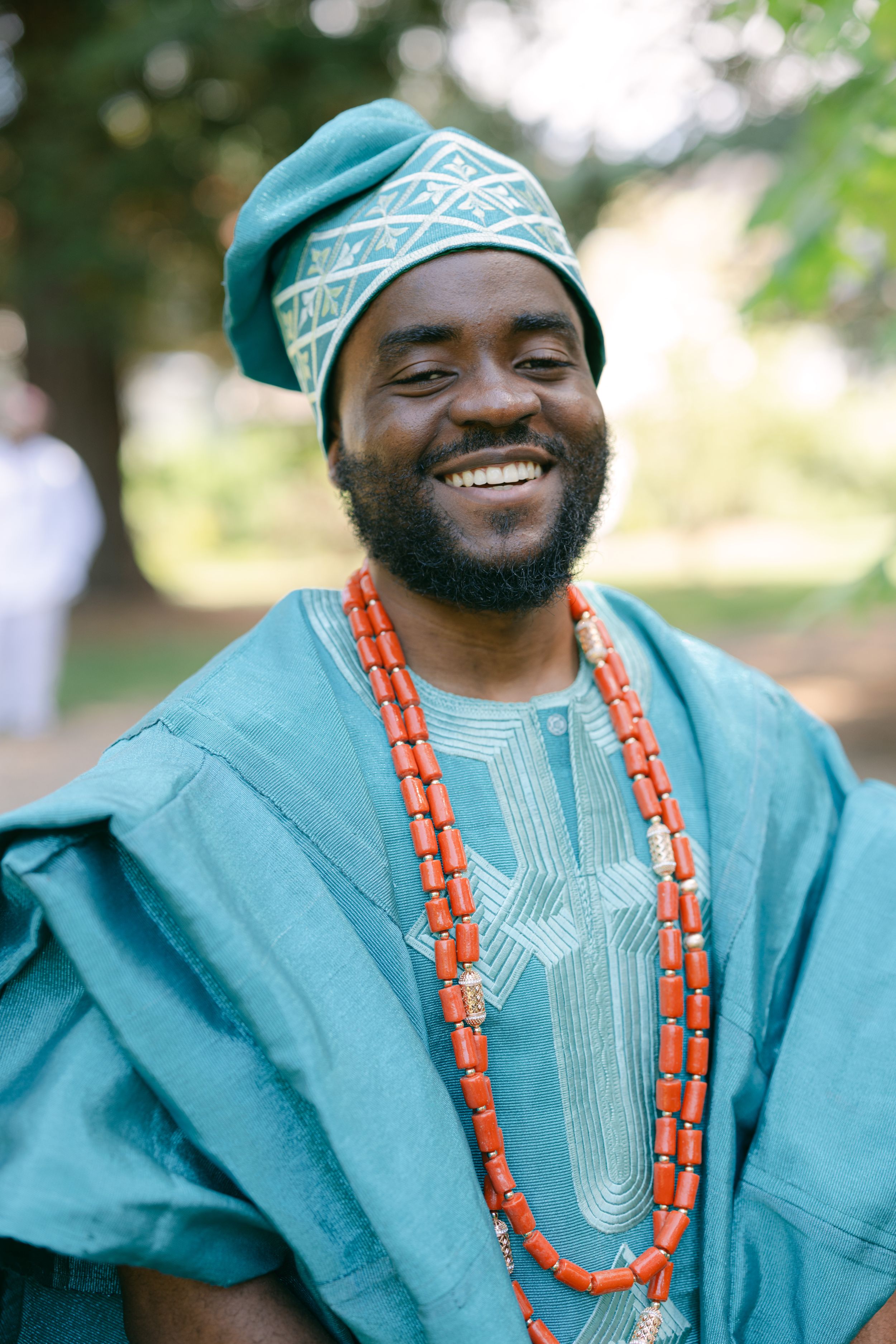 Traditional African fashion featuring turquoise agbada outfit with orange beaded necklace photographed outdoors.