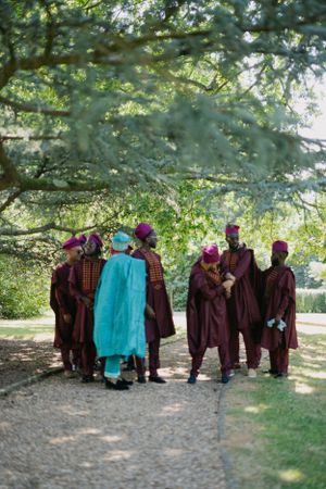 Wedding party members in burgundy robes and turquoise attire gather under tree branches.