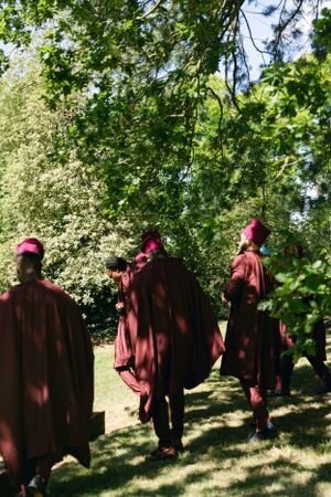 Three figures in flowing burgundy robes and caps walking through a sunlit garden setting.