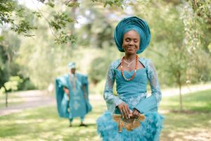 Person in turquoise traditional dress and headwrap standing in a sunny garden setting.