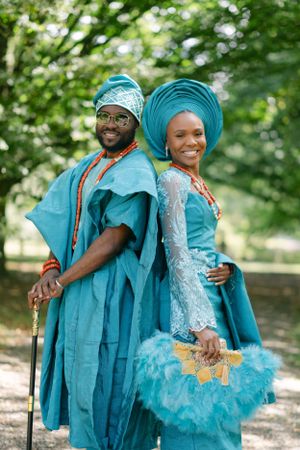 A couple in matching blue Nigerian outfits smile and pose playfully in a park setting.