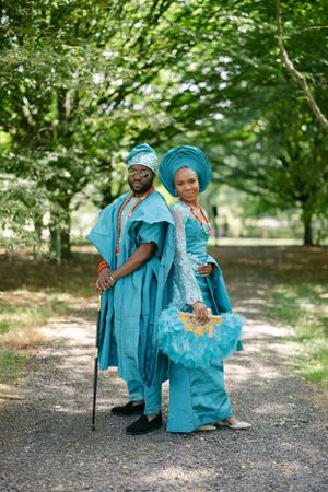 A couple in traditional Nigerian dress pose back-to-back on a forest path.