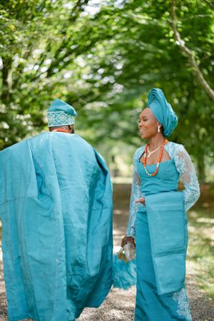 A couple in traditional blue Nigerian formal wear pose on a tree-lined path.