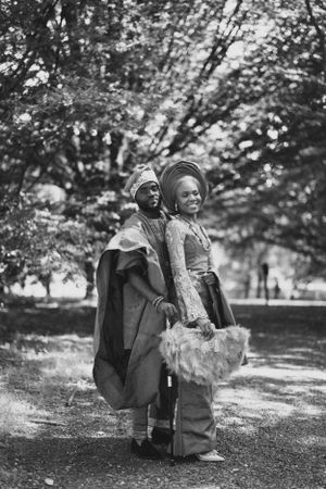 A black and white portrait of a couple in traditional African formal wear standing together outdoors.