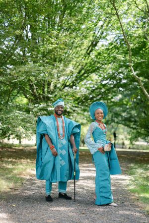 Couple in coordinated turquoise Nigerian formal wear walking along a tree-lined path.
