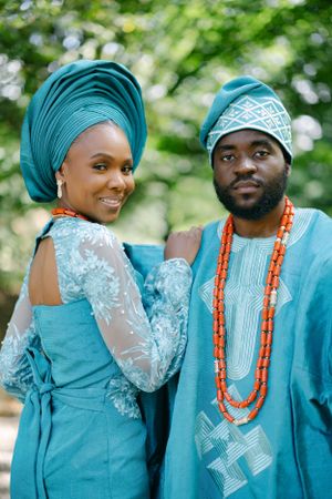 A couple in coordinated turquoise Nigerian wedding attire pose against a leafy backdrop.