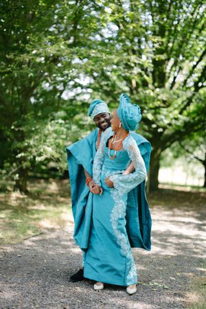 Couple in matching turquoise traditional African wedding attire posing on a tree-lined path.