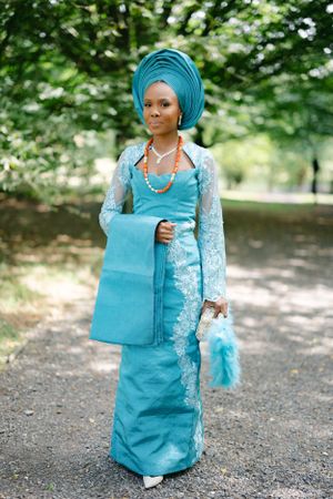 Woman in turquoise Nigerian gele and lace dress with coral necklace standing on garden path.