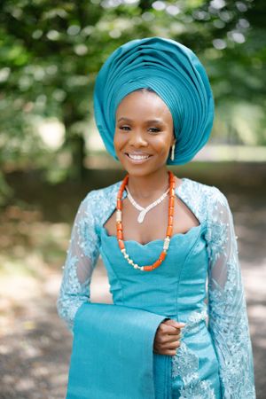 Traditional bride wearing turquoise gele and lace dress with coral beaded necklace in garden setting.