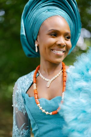 Woman in turquoise gele and dress smiling while wearing coral and pearl beaded necklace.