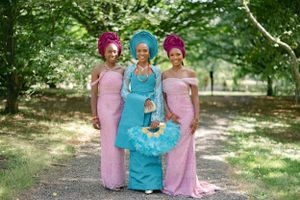 Three women in formal attire wearing pink and blue dresses with matching gele headwraps in outdoor setting.