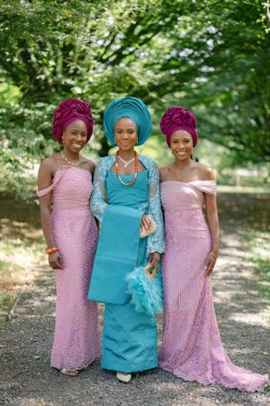 Three women pose in pink and turquoise formal dresses with matching gele headwraps on a garden path.
