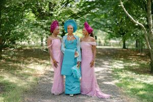 Three women in traditional African attire pose outdoors wearing pink and turquoise dresses with matching headwraps.