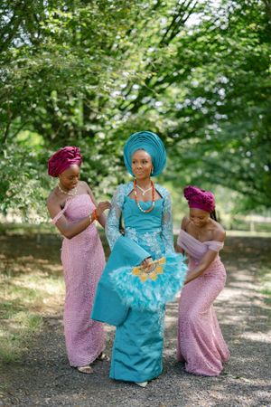 Three women in traditional African dress pose playfully on garden path wearing pink and turquoise outfits.