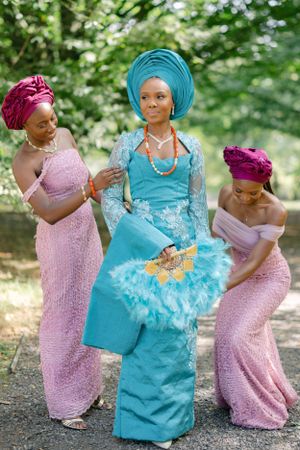 Three women in traditional African attire share a moment while wearing coordinated pink and turquoise outfits.