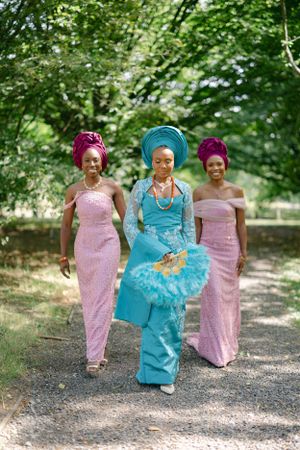 Three women in traditional Nigerian attire walk together, wearing pink and turquoise dresses with matching head wraps.