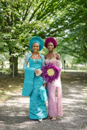 Two women in formal attire pose together on a tree-lined path, holding vibrant purple flowers.