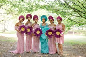A wedding party poses together wearing pink dresses and traditional head wraps with purple floral bouquets.
