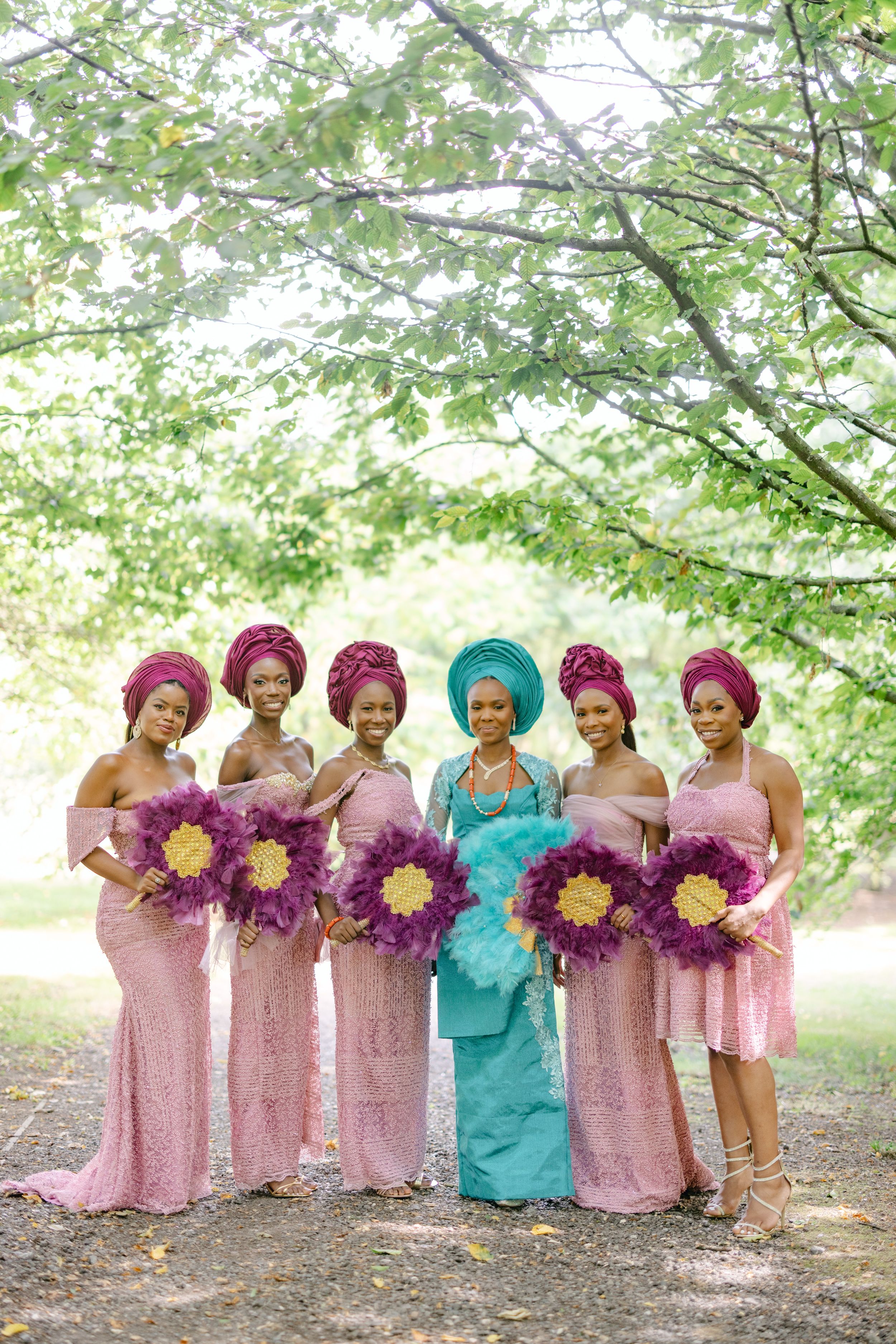 A bridal party poses outdoors wearing pink dresses and bright head wraps with purple floral bouquets.
