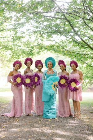 Wedding group in pink gowns and traditional head wraps holds vibrant purple and yellow bouquets outdoors.