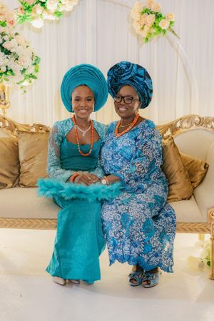 Two women wearing vibrant turquoise and blue traditional Nigerian attire pose on an elegant gold couch.